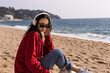Stylish teenager model wearing hoodie and headphones listening dj music, relaxing on the beach. Young teen girl enjoy sunny day outdoors and listen to music or audiobook, learn new language.