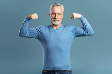 Strong gray haired senior man showing muscles isolated on blue background. Handsome active 60 years old muscular male looking at camera. Gym workout, sport, healthy lifestyle concept