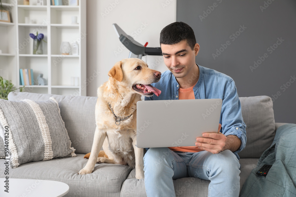 Young man with cute Labrador dog using laptop on sofa at home