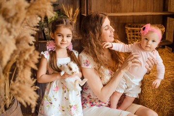 A mother holds six-month-old infant upright in her arms, while her second daughter, wearing a summer dress and braided hair, stands beside holding a rabbit in hands