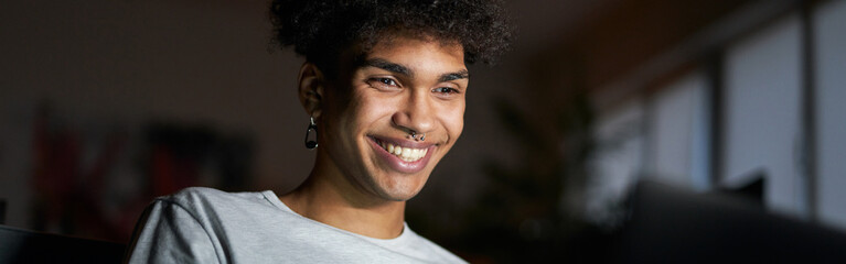 Wall Mural - Night portrait of young smiling guy working on laptop in dark room, sitting on a couch at home