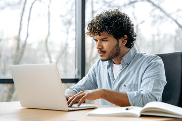 Puzzled frustrated indian or arabian curly man, financial manager, company ceo, sitting at a desk in the office, looking at a laptop screen in confusion, experiencing misunderstanding, anxiety, stress