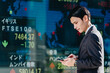 世界の株価一覧が表示された電光掲示板の前でスマホアプリで取引を注文する男性サラリーマン