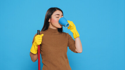 Wall Mural - Cheerful housekepper enjoying cup of coffee while cleaning house with broom, standing in studio over blue background. Maid using protective equipment and sanitary practices to keep customers safe.