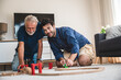 © chokniti - Closeup of a young man and elderly father playing with toy train set at home by sitting on floor in the living room and having fun and enjoying