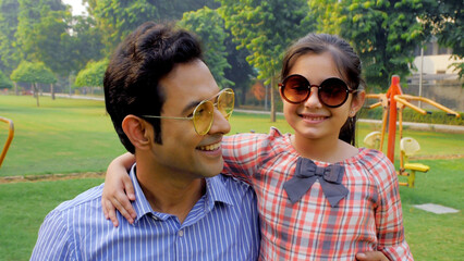 Wall Mural - Man and his happy child wearing sunglasses enjoying in a park during summers . Medium shot of cheerful father holding his daughter in arms while standing near a green area