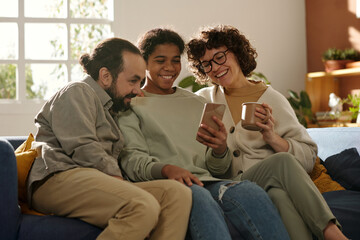 Wall Mural - Teenage girl watching funny video on smartphone with her forest people, they sitting on sofa and laughing during leisure time at home