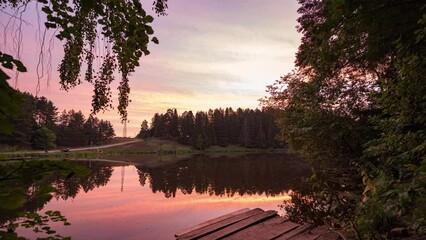 Wall Mural - Time-lapse sunrise over beautiful lake. Water surface in fast motion. Warm sky with rolling clouds. Trees silhouette woodland on horizon. Early morning shot in timelapse. Bright sun rises over skyline