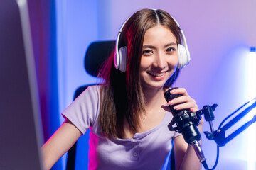 Professional gamer. Young asian pretty woman sitting on chair with computer pc in living room. Happy female Professional Streamer chinese wearing headphone playing game online in dark room neon light