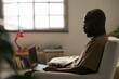 © pressmaster - African AMerican man sitting on armchair in the room and working online using his laptop