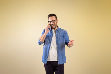 Wall Mural - Young male entrepreneur sharing good news over smart phone and laughing cheerfully on isolated beige background. Businessman dressed in denim shirt talking over cellphone