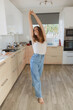 © master1305 - Full-length image of young smiling, positive, relaxed woman in casual clothes standing at home in kitchen on daytime. Stylish, cozy interior. Concept of home life, emotions, vacation, weekends