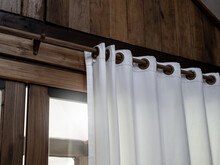 Close-up Of White Wall Curtain On Wooden Curtain Rail Bar Decorated On The Wood Plank Wall Of Tropical Gable Building Near The Sliding Glass Door Inside Room With Sunlight From Outside.