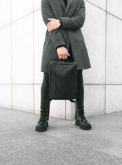 Vertical shot of a young male wearing a gray coat and holding a black backpack on the street