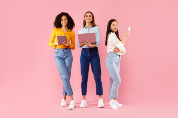 Three diverse ladies using different gadgets, networking online, standing on pink background, full length