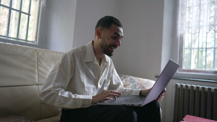 One excited happy young Middle Eastern man reacting positively to successful message online while sitting on couch in front of laptop computer