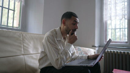 One excited happy young Middle Eastern man reacting positively to successful message online while sitting on couch in front of laptop computer