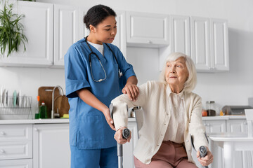 Wall Mural - multiracial nurse in uniform helping senior woman using crutches to stand up in modern apartment.