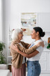 © LIGHTFIELD STUDIOS - side view of happy multiracial social worker hugging with senior woman in living room.