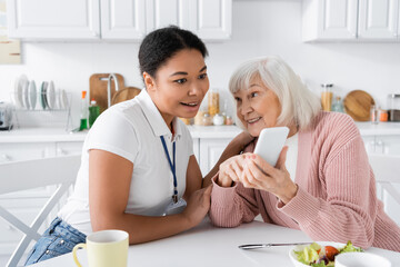 Wall Mural - happy senior woman showing smartphone to amazed multiracial social worker at home.