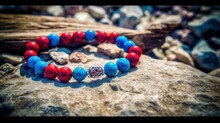  A Red, White And Blue Beaded Bracelet Sitting On A Rock Next To A Tree Trunk And A Rock With A Silver Charm On It.  Generative Ai
