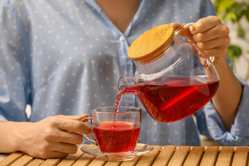 Woman pouring delicious hibiscus tea into cup at wooden table, closeup