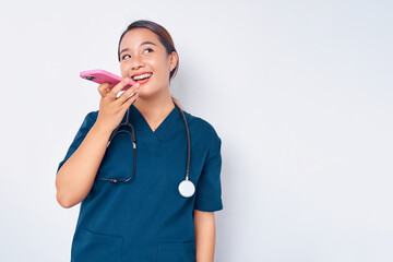 Wall Mural - Beautiful smiling young Asian woman professional nurse working wearing a blue uniform talking on mobile phone in loudspeaker mode isolated on white background. Healthcare medicine concept