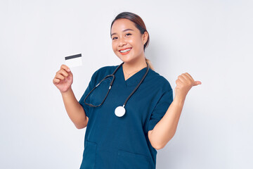 Wall Mural - Beautiful smiling young Asian woman professional nurse working wearing blue uniform holding credit bank card and pointing thumb at copy space isolated on white background. Healthcare medicine concept