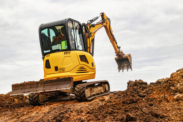Fototapete - Mini excavator at the construction site on the edge of a pit against a cloudy blue sky. Compact construction equipment for earthworks. An indispensable assistant for earthworks.