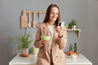 © sementsova321 - Joyful positive woman with brown hair wearing beige jacket standing in home kitchen interior having breakfast and using mobile phone checking social networks in morning.