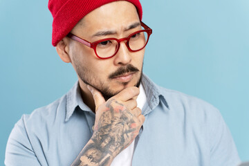 Wall Mural - Closeup portrait of handsome pensive asian man wearing red hat, stylish eyeglasses isolated on blue background. Serious Korean hipster brainstorming, planning startup indoors, studio shot