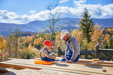 Wall Mural - Father with toddler son building wooden frame house. Boy helping his daddy, hammering nail into plank on construction site, wearing helmet and blue overalls on sunny day. Carpentry and family concept.