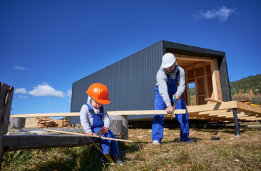 Wall Mural - Father with toddler son building wooden frame house. Boy helping his daddy, using hand saw to cut boards on construction site, wearing helmet, blue overalls on sunny day. Carpentry and family concept.