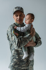 Canvas Print - American army soldier in military uniform with cap holding infant son in arms isolated on grey.