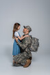 happy serviceman in military uniform and backpack hugging cheerful daughter on his homecoming on grey background.