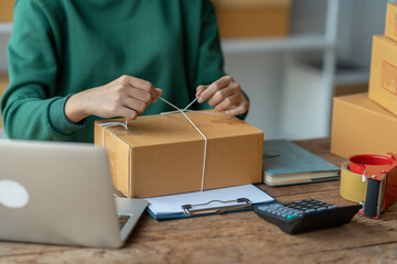 Asian businesswomen tie boxes with ropes in preparation for delivery to customers. Young Asian small business owners use computers. Shipping boxes online market SME e-commerce telemarketing concept.