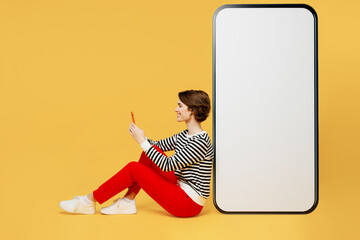 Full body sideways happy young woman wear casual black and white shirt sit big huge blank screen mobile cell phone with workspace mockup area using smartphone isolated on plain yellow color background