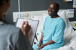 © AnnaStills - Smiling elderly patient sitting on bed and talking to nurse about his health condition while she making noted in medical card
