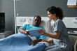 © AnnaStills - Young nurse showing medical history on digital tablet to elderly patient while he lying on bed in ward