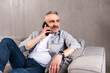 © F8  \ Suport Ukraine - Successful and smiling middle-aged man in white shirt talking by the phone at home. Sitting on the couch and looking in front of him.