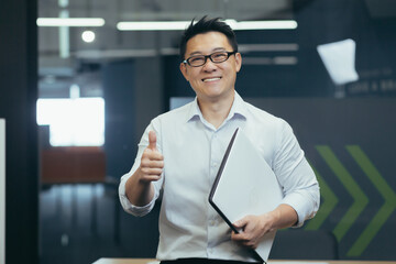 portrait of a successful young asian male programmer standing in a modern office with a notebook in 