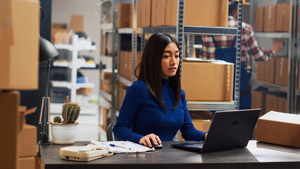 Wall Mural - Young adult working on retail stock orders in depot, checking distribution and shipment on computer. Female owner checking merchandise on storage room racks, business management. Tripod shot.