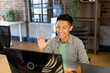 © Wavebreak Media - Smiling asian male creative at computer making video call, waving in casual office