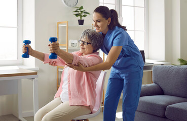 Wall Mural - Young nurse or physiotherapist in scrubs helping a happy retired old woman do fitness exercises with light weight dumbbells at home. Concept of physiotherapy for seniors