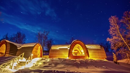 A starry sky in the blue sky above a winter landscape with dome-shaped wooden houses. Time Lapse