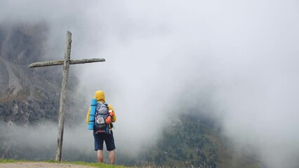Wall Mural - young man hiker standing near wooden cross on mountain peak with cloudy sky and fog. yellow hood jac