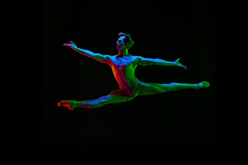 Portrait of young handsome, muscle men, ballet dancer in pastel color clothes jumping over dark background with neon light. Contemporary dance style. Split jump