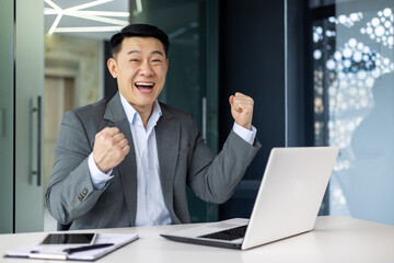 Wall Mural - Portrait of successful asian boss, businessman celebrating results achievement, man in business suit smiling and looking at camera holding hands up triumph gesture, mature investor inside office.