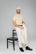 Full length of trendy homosexual man in hat holding jacket near chair on grey background.