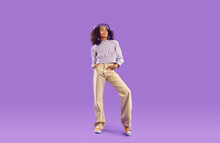 Kid Model In Stylish Fit Posing In Studio. Full Body Length Beautiful African Girl Wearing Beret Hat, Blouse And Beige Wide Leg Jeans Standing On Purple Background. Clothing, Childrens Fashion Concept